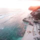 Aerial view of Melasti Beach Ungasan, Bali, showcasing turquoise waters, white sand, and dramatic limestone cliffs at sunset.