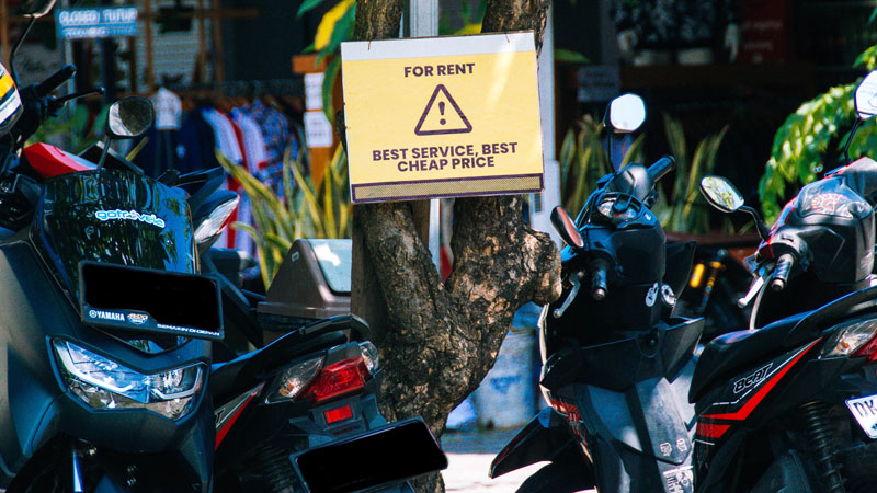 Scooters available for rent near Legian Beach with a sign advertising 'Best Service, Best Cheap Price'.