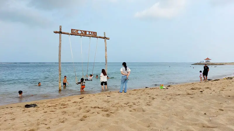 Families and children enjoying swings and calm waters at Sanur Beach Bali, with a traditional Balinese gazebo in the background.