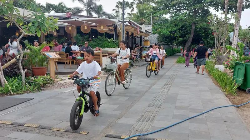 Families cycling along the Sanur Promenade with cafés in the background.