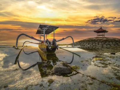 Sunrise over a jukung boat on Sanur Beach, Bali, with tranquil waters and a traditional gazebo in the background.